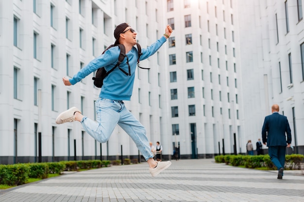 Il giovane ragazzo alla moda in vestiti blu salta vicino a un edificio moderno bianco