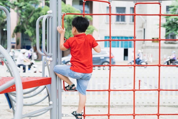 Il giovane ragazzo asiatico si arrampica sulla recinzione di corda rossa e sulla barra grigia con la mano per esercitarsi nel parco giochi all'aperto sotto il grande albero.