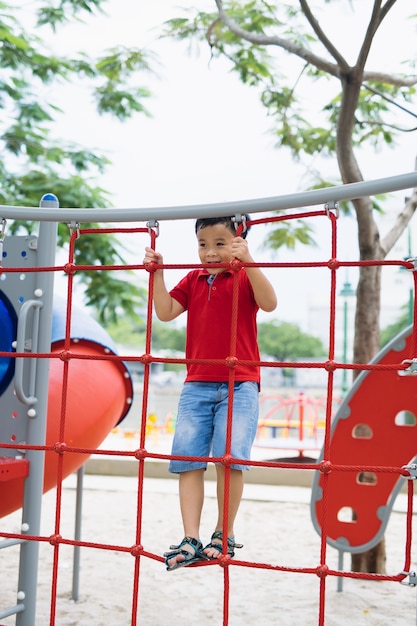Il giovane ragazzo asiatico si arrampica sulla recinzione di corda rossa e sulla barra grigia con la mano per esercitarsi nel parco giochi all'aperto sotto il grande albero.