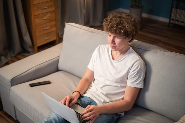 Il giovane ragazzo con i capelli ricci si siede sul divano con un computer portatile e guarda la tv