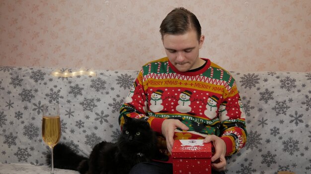 Il giovane ragazzo di vacanze di Capodanno apre e avvolge un regalo, champagne, regali di Capodanno, decorazioni di Capodanno e buon umore e un gatto nero