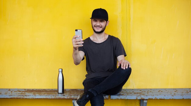 Il giovane ragazzo sorridente è seduto dietro il giallo e sta prendendo selfie con lo smartphone vicino alla sua bottiglia termica per l'acqua.