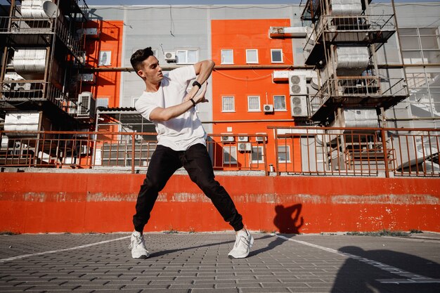 Il giovane ragazzo vestito in jeans e t-shirt sta ballando la danza moderna in strada sullo sfondo degli edifici urbani nella calda giornata.