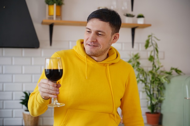 Il giovane rilassato posa con un bicchiere di vino rosso in piedi al tavolo della cucina Ragazzo adulto che riposa con l'alcol in cucina
