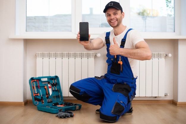 Il giovane riparatore professionista in uniforme speciale con gli strumenti sta installando i radiatori e il termostato nella stanza domestica