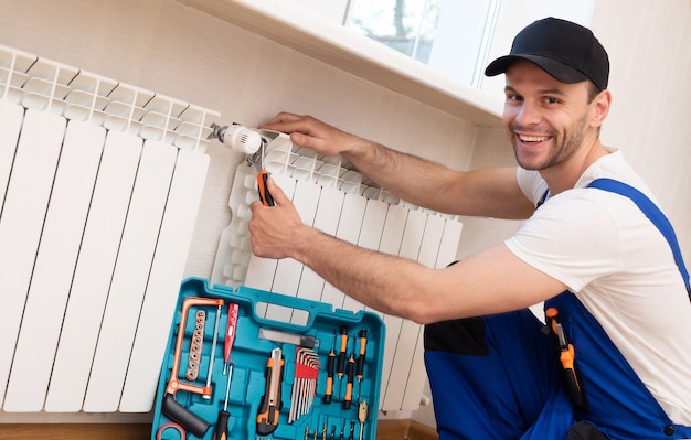 Il giovane riparatore professionista in uniforme speciale con gli strumenti sta installando i radiatori e il termostato nella stanza domestica