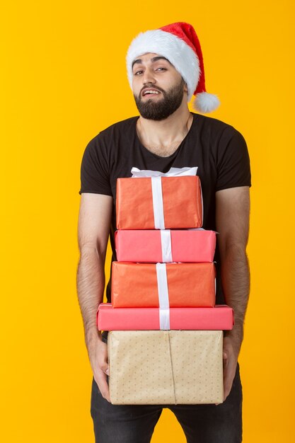 Il giovane scontento con la barba in un cappello di Babbo Natale tiene cinque scatole regalo in posa su un muro giallo. Concetto di regali e auguri per il Natale