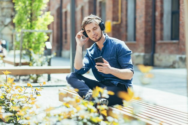Il giovane seduto su una panchina fuori dall'ufficio ad ascoltare musica gode di un'applicazione mobile per uno smartphone con cuffie grandi