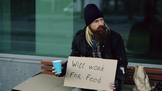 Il giovane senzatetto chiede soldi scuotendo la tazza per prestare attenzione alle persone che camminano vicino