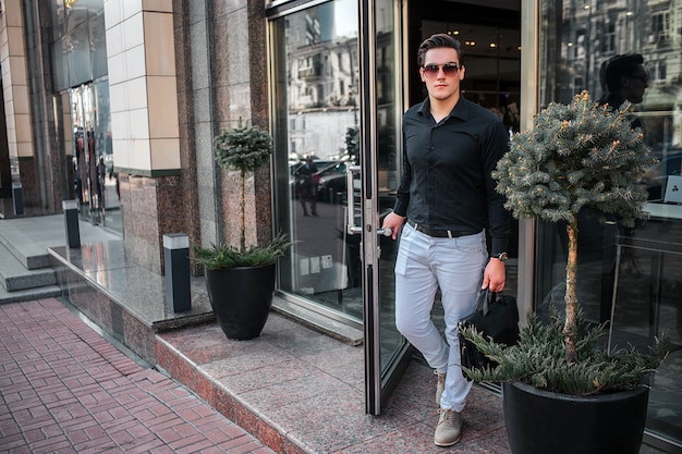 Il giovane serio cammina fuori dell'edificio. Ha aperto la porta. Guy tiene in mano una borsa nera.