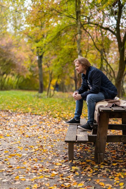 Il giovane si siede all'aperto nel periodo autunnale. Maschio seduto su una panchina in un parco autunnale. Bel ragazzo felice su una panca di legno