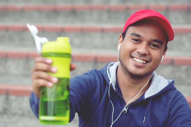 Il giovane si siede ascolta musica felicemente rompe l'acqua potabile dopo aver eseguito l'allenamento di jogging. Maschio sano assetato con acqua potabile del corpo in forma.