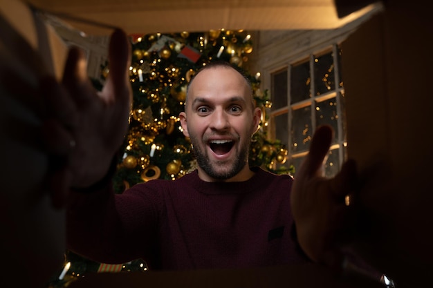 Il giovane sorridente che apre un regalo di Natale a casa un uomo guarda e ride si mette le mani dentro