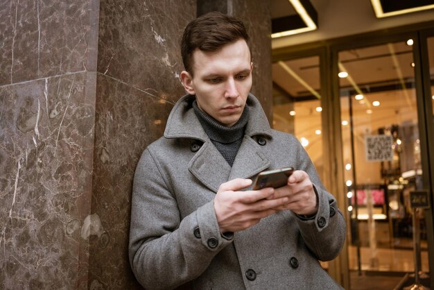 Il giovane sorridente in cappotto grigio sta tenendo i messaggi di testo del rotolo nel suo cellulare