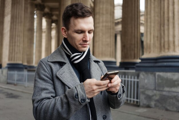 Il giovane sorridente in cappotto grigio sta tenendo i messaggi di testo del rotolo nel suo cellulare