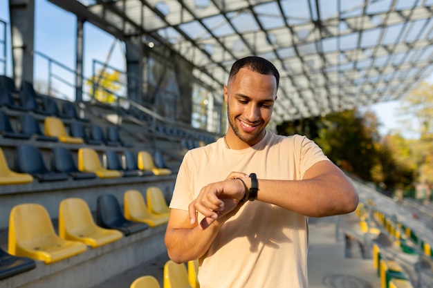 Il giovane sportivo maschio latinoamericano si trova allo stadio e guarda il braccialetto fitness