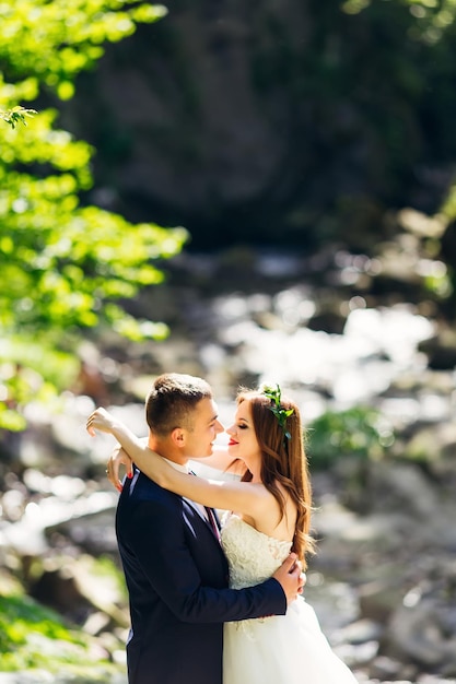 Il giovane sposo in abito blu si abbraccia in un parco con piante verdi con una bella sposa dai capelli ricci