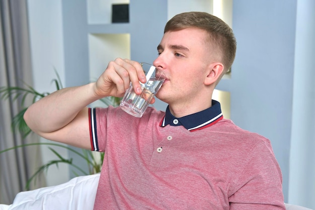 Il giovane sta bevendo acqua fresca pura dal vetro a casa nel soggiorno