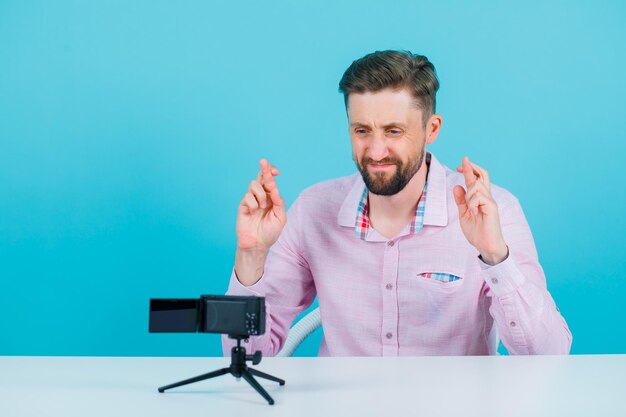 Il giovane sta posando alla sua mini macchina fotografica incrociando le dita su sfondo blu