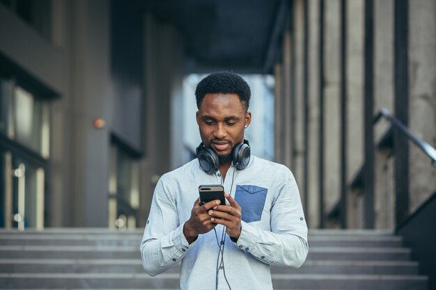 Il giovane studente afroamericano ascolta il podcast utilizzando l'app sul telefono e un grande auricolare, in abiti casual del giorno