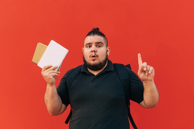 Il giovane studente maschio grasso sorpreso con la barba sta su fondo rosso con il libro