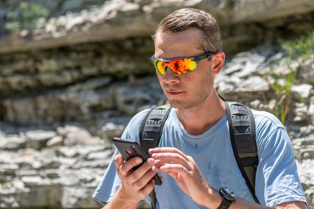 Il giovane turista maschio in occhiali sportivi si siede con un telefono cellulare in mano in montagna