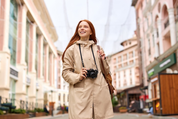 Il giovane turista sorridente della donna cammina intorno alla città in primavera, tiene la retro macchina fotografica. Donna rossa in viaggio, in abito casual in stile urbano. Ritratto di signora caucasica all'aperto, da sola