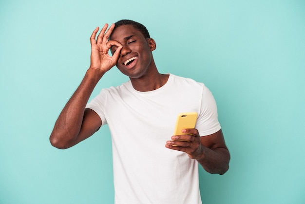 Il giovane uomo afroamericano che tiene un telefono cellulare isolato su fondo blu ha eccitato mantenendo il gesto giusto sull'occhio.