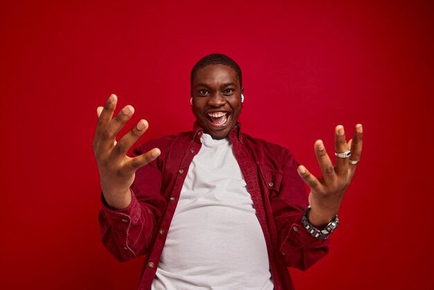 Il giovane uomo afroamericano si rallegra con le mani alla macchina fotografica isolata sul ritratto in studio di sfondo rosso