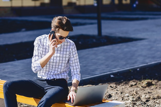 Il giovane uomo alla moda lavora al computer portatile al parco il giorno soleggiato