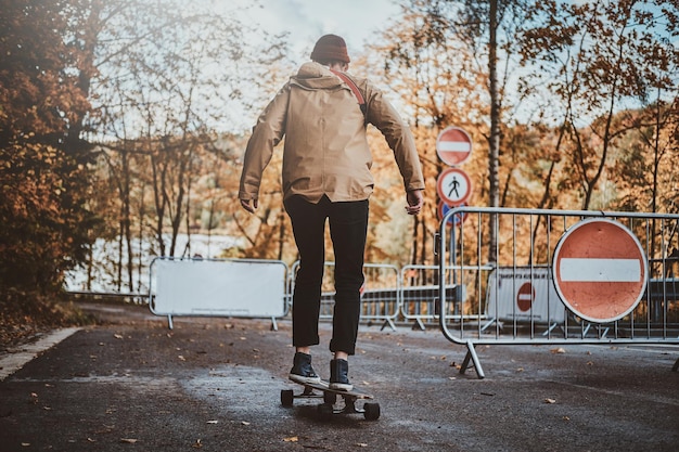 Il giovane uomo alla moda sta cavalcando il suo longboard al parco autunnale circondato da alberi gialli.