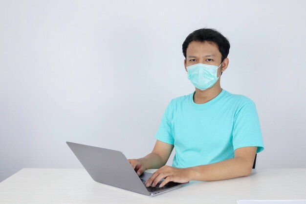 Il giovane uomo asiatico che indossa una maschera medica è serio e si concentra quando lavora su un laptop sul tavolo Uomo indonesiano che indossa una camicia blu