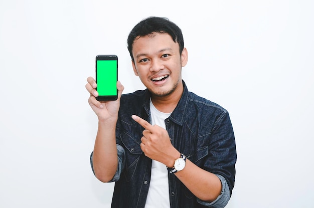 Il giovane uomo asiatico è in piedi e sorridente puntando sullo schermo verde sullo schermo dello smartphone su sfondo bianco