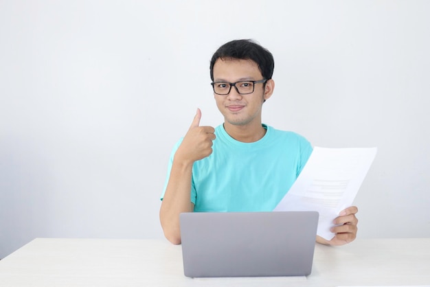 Il giovane uomo asiatico è sorridente e felice quando si lavora su un laptop e si documenta con il segno della mano ok Uomo indonesiano che indossa una camicia blu