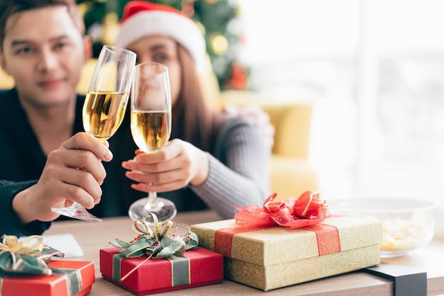 Il giovane uomo asiatico felice sorride e ride alle spalle della sua ragazza che indossa un cappello di Babbo Natale mentre beve champagne a casa con un albero di Natale sullo sfondo