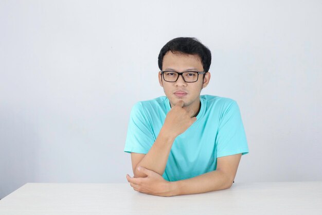 Il giovane uomo asiatico indossa una camicia blu e occhiali con la faccia seria che guarda sulla fotocamera e tiene il mento sul tavolo