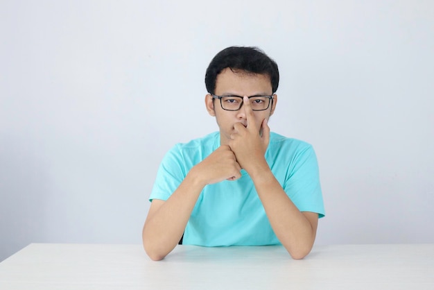 Il giovane uomo asiatico indossa una camicia blu e occhiali con la faccia seria che guarda sulla fotocamera e tiene il mento sul tavolo