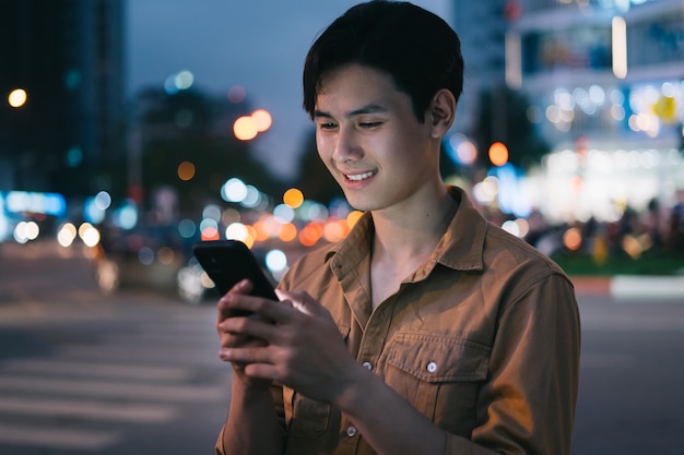 Il giovane uomo asiatico sta usando il suo telefono mentre cammina per strada di notte