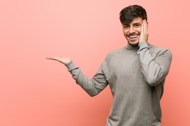 Il giovane uomo astuto dello studente tiene lo spazio della copia su una palma, tiene la mano sulla guancia. Stupito e felice.