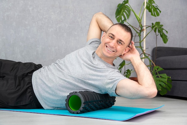 Il giovane uomo atletico sorridente fa un esercizio di rilascio miofasciale sul tappetino da yoga con rullo di schiuma e guarda la telecamera. Esercizio MFR.