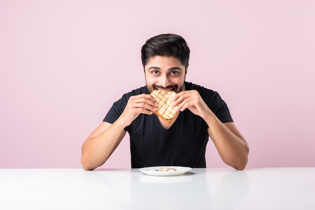 Il giovane uomo barbuto asiatico indiano mangia panino di pane mentre è seduto in cucina o al tavolo da pranzo. Mostrare o presentare