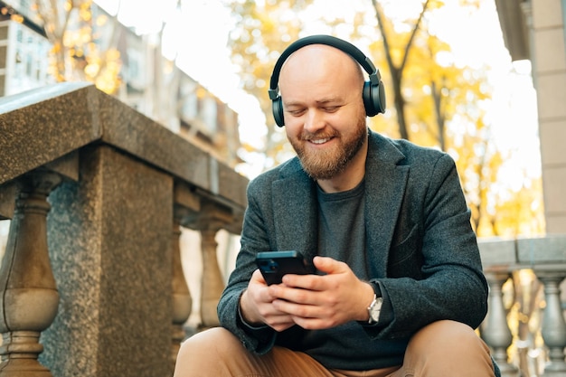 Il giovane uomo barbuto calvo gode di un podcast musicale o di un audiolibro mentre usa il telefono