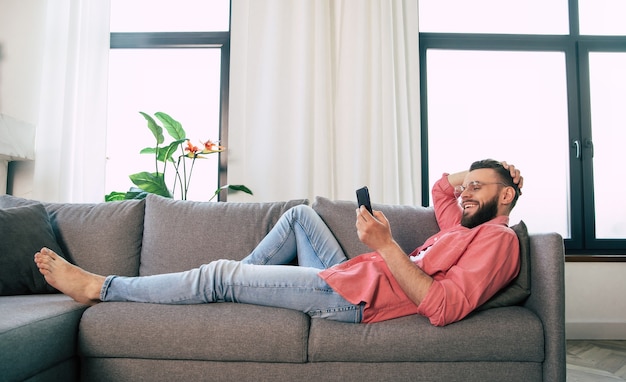 Il giovane uomo barbuto felice bello con gli occhiali sta usando lo smartphone per navigare in rete, digitare e chattare, giocare o lavorare con alcune app mentre ci si rilassa sul divano di casa