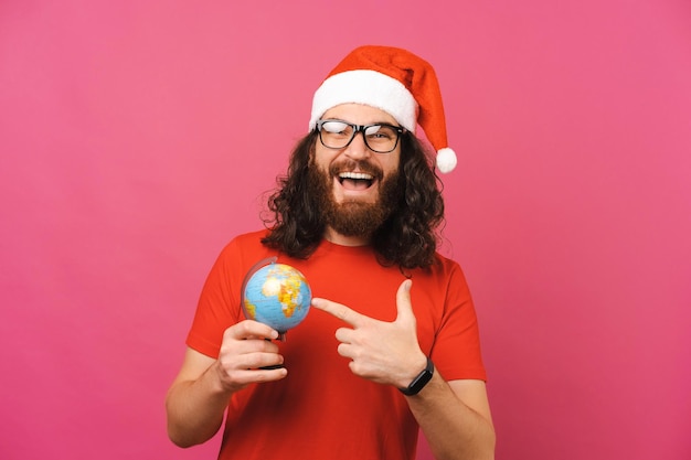 Il giovane uomo barbuto riccio che indossa il cappello di Natale sta puntando sul globo terrestre