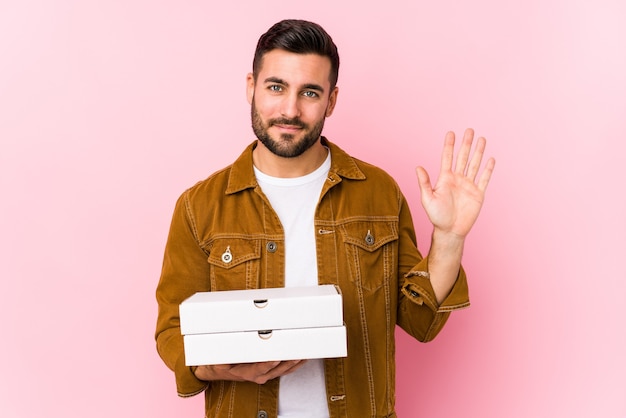 Il giovane uomo bello che tiene le pizze ha isolato sorridente allegro che mostra il numero cinque con le dita.