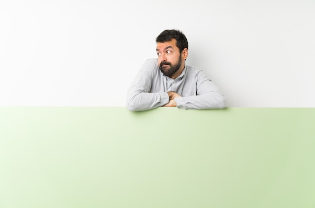 Il giovane uomo bello con la barba che tiene un grande cartello vuoto verde che fa i dubbi fa gesto mentre solleva le spalle