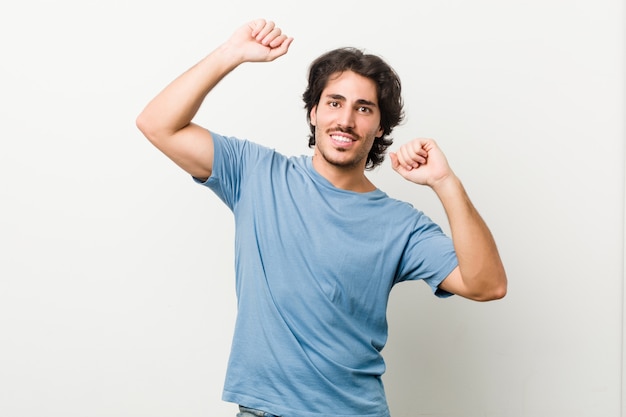 Il giovane uomo bello contro una parete bianca che celebra un giorno speciale, salta e alza le braccia con energia.