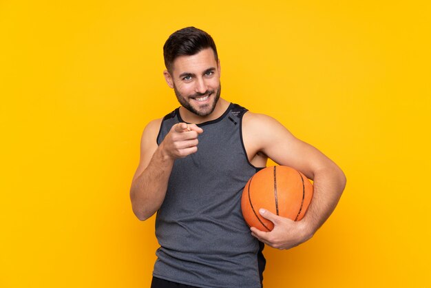 Il giovane uomo bello del giocatore di pallacanestro sopra la parete bianca isolata indica il dito con un'espressione sicura