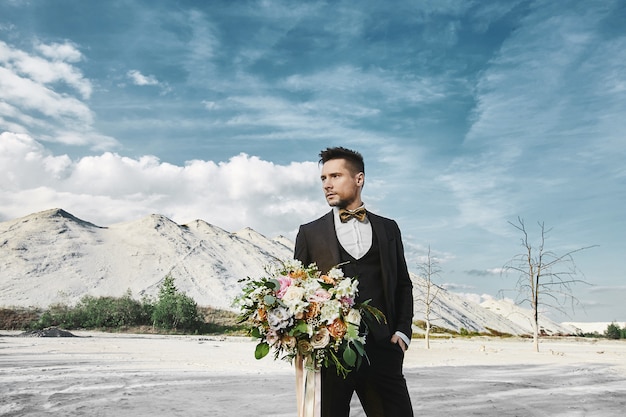 Il giovane uomo bello ed elegante in vestito alla moda e farfallino sta tenendo un grande mazzo di fiori e guarda da parte il deserto