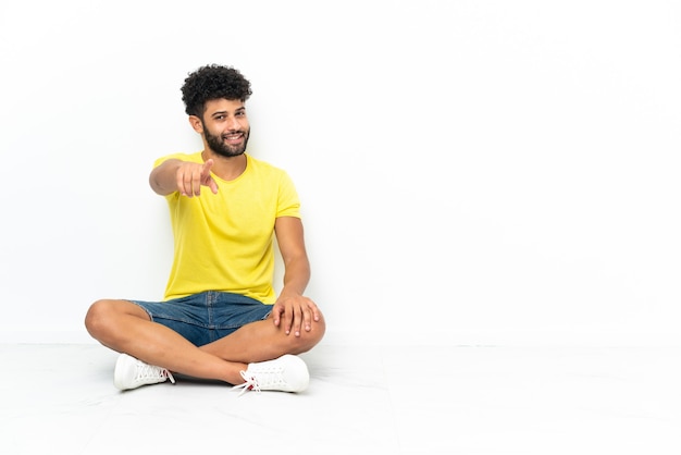 Il giovane uomo bello marocchino seduto sul pavimento su sfondo isolato punta il dito contro di te con un'espressione fiduciosa